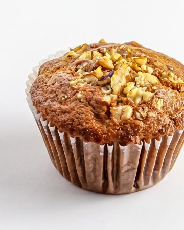 Banana nut muffin with chopped walnuts on top in a brown paper liner against white background.