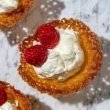 Crispy golden lace cookies filled with whipped cream and topped with fresh red raspberries on a marble surface.