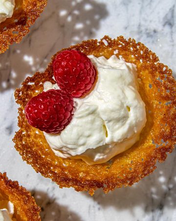 Crispy golden lace cookies filled with whipped cream and topped with fresh red raspberries on a marble surface.