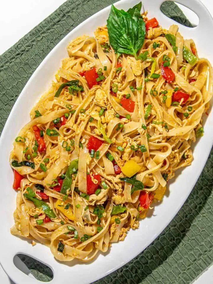 Stir-fried flat rice noodles with scrambled eggs, red and yellow bell peppers, green onions, and a basil leaf garnish on a white oval platter.