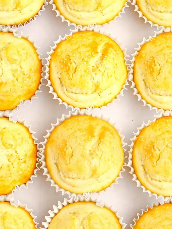 Close-up of several golden-brown muffins in white paper liners arranged in a grid pattern on a light surface.