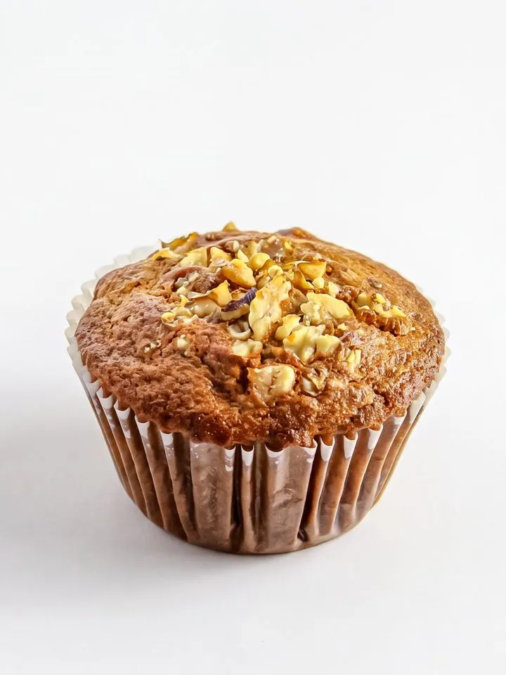 Banana nut muffin topped with chopped walnuts in a white paper liner on a white background.