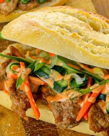 Grilled beef banh mi sandwich with pickled carrots, cucumber ribbons, scallions, and spicy mayo on a crusty baguette on a wooden board.