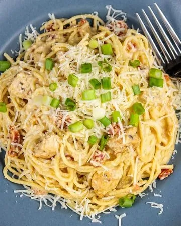 Spaghetti pasta with creamy sauce, chunks of chicken, tomatoes, shredded cheese, and chopped green onions on a blue plate with a fork.
