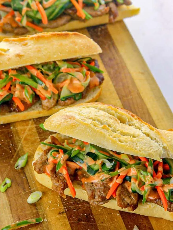 Three banh mi sandwiches with grilled meat, pickled vegetables, and spicy mayo on a wooden cutting board.
